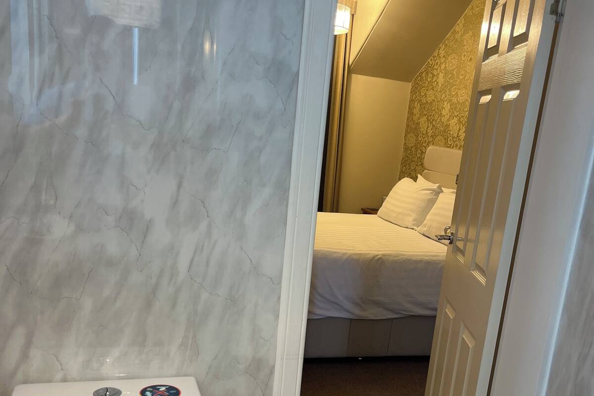 Une salle de bain comprenant des toilettes, un espace douche et un lit visible à travers une porte ouverte.