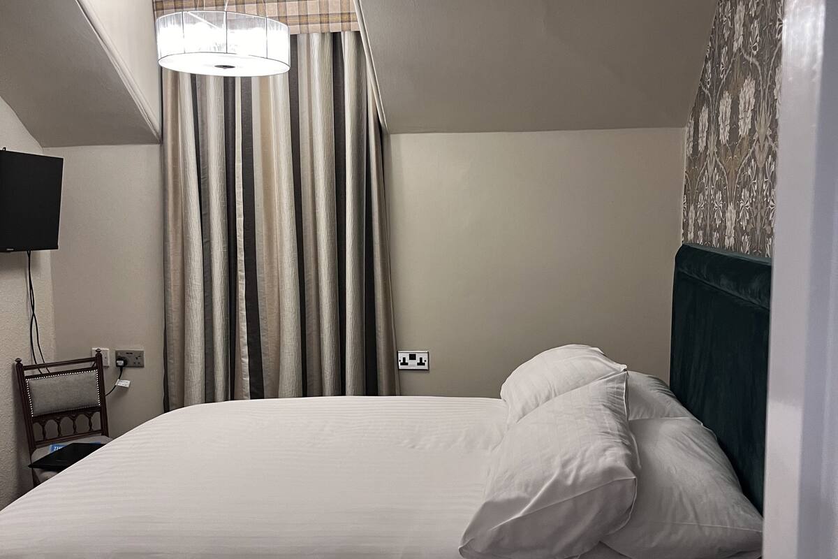Un lit bien fait, avec du linge de lit blanc, une table de chevet en bois avec une télécommande, un téléviseur fixé au mur et un mur décoré d’un motif.