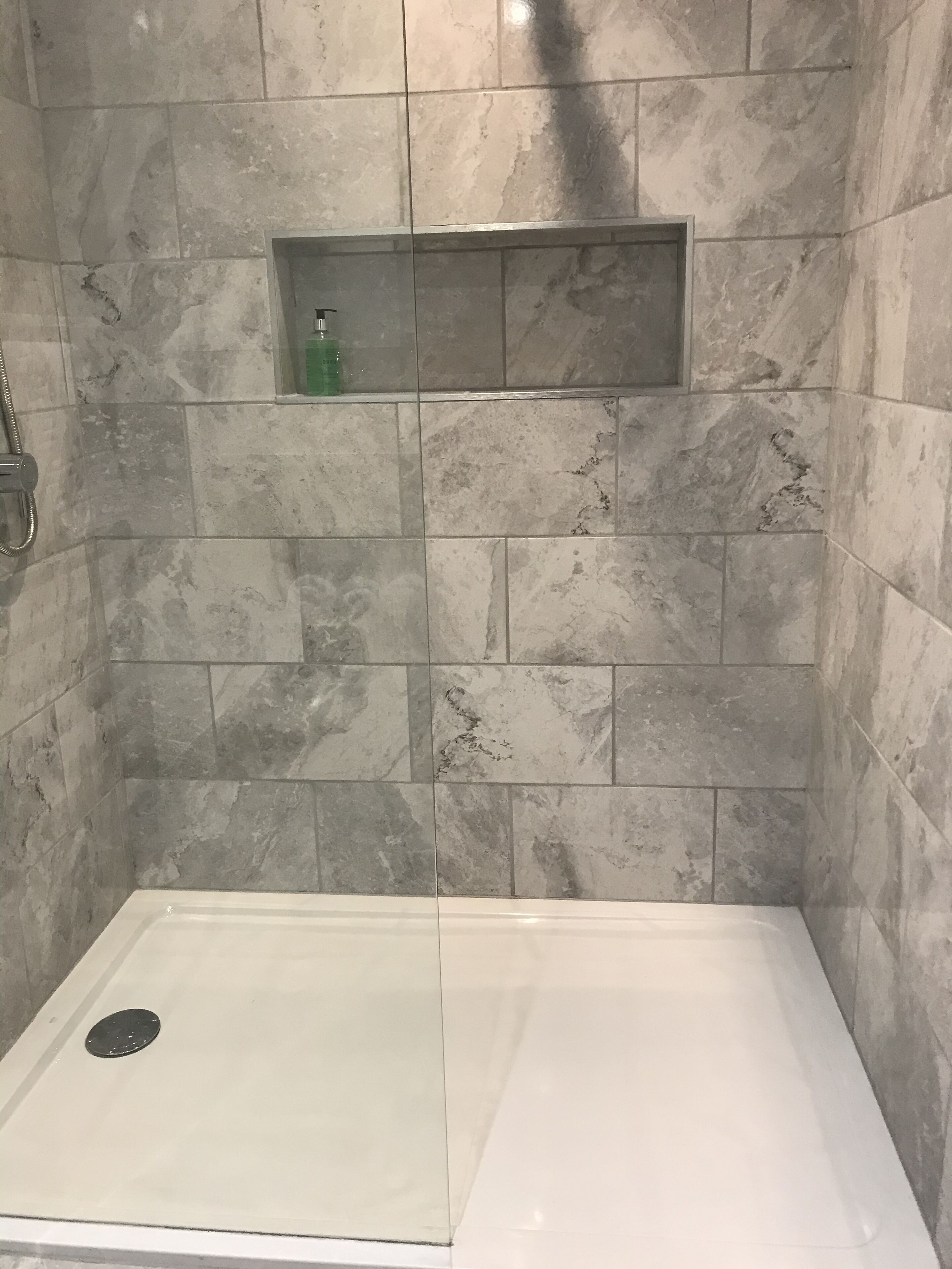A shower area with a glass door, a built-in shelf with a bottle, and a drain.