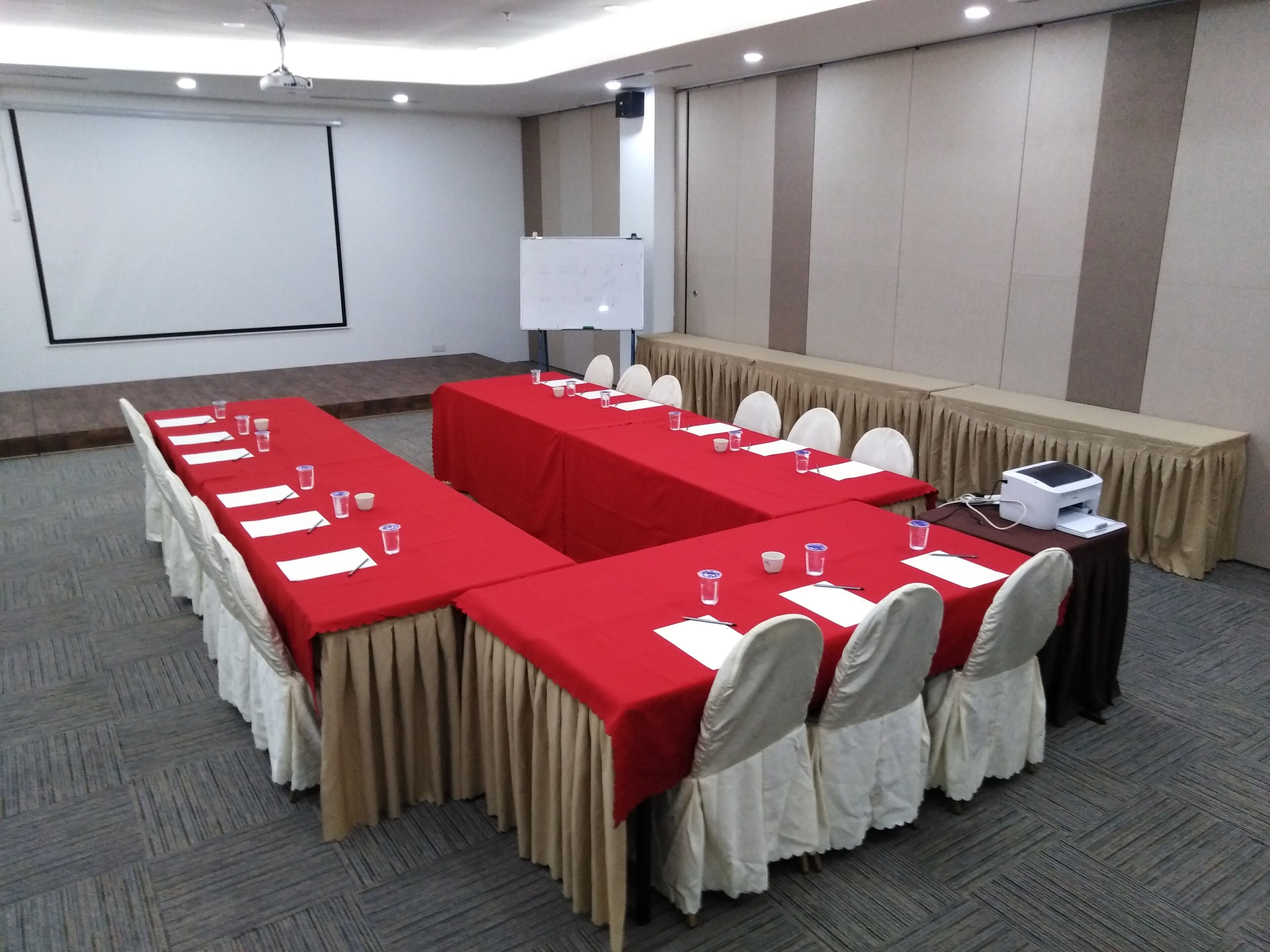 Uma sala de conferências com mesas grandes, cadeiras brancas e uma tela de projeção.