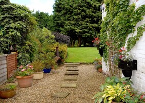 A garden pathway leading to a lawn, surrounded by various potted plants and a rustic chimney.