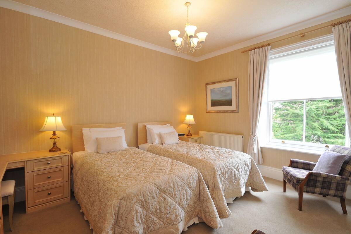 Een hotelkamer met twee bedden, een stoel, een bureau met lamp en een raam met gordijnen.