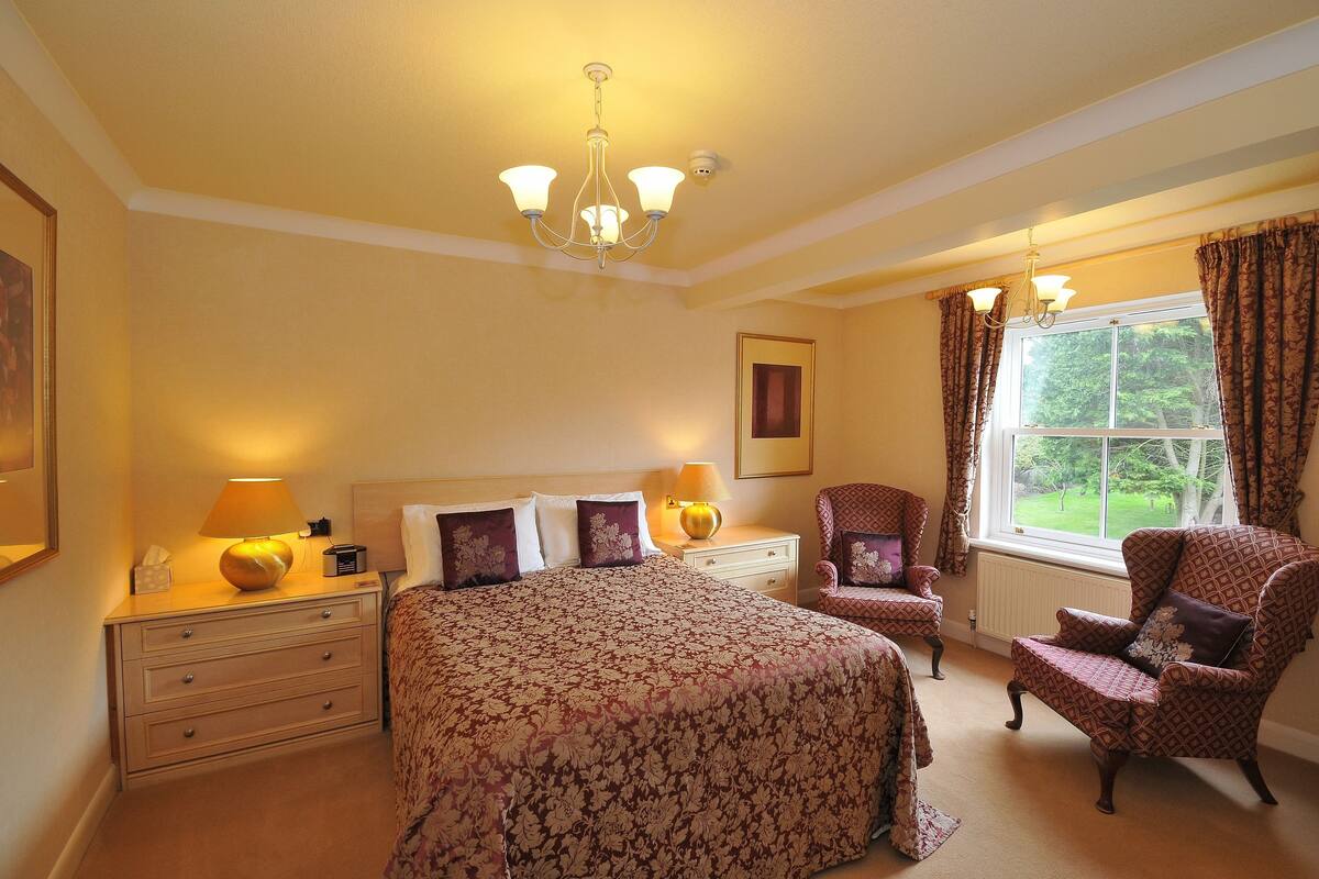 Een slaapkamer met een groot bed, twee nachtkastjes met lampen, een stoel, een raam met gordijnen en een kroonluchter.