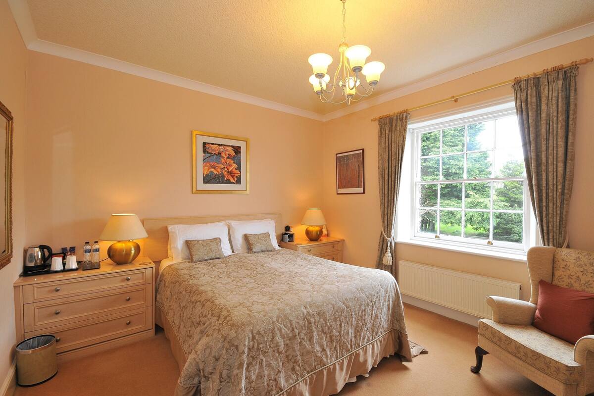 Een goed verlichte slaapkamer met een groot bed, nachtlampjes, een raam met gordijnen en een stoel.