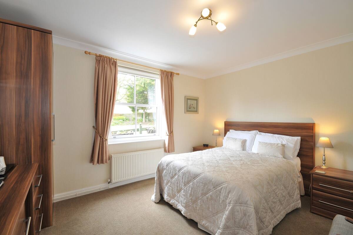 Een slaapkamer met een groot bed, een houten garderobe, een nachtkastje met lamp, een raam met gordijnen en een radiator.