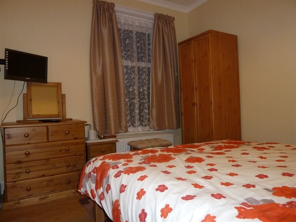 A bedroom with a wooden dresser, a bed with a floral bedspread, a wooden wardrobe, and a television on a stand.
