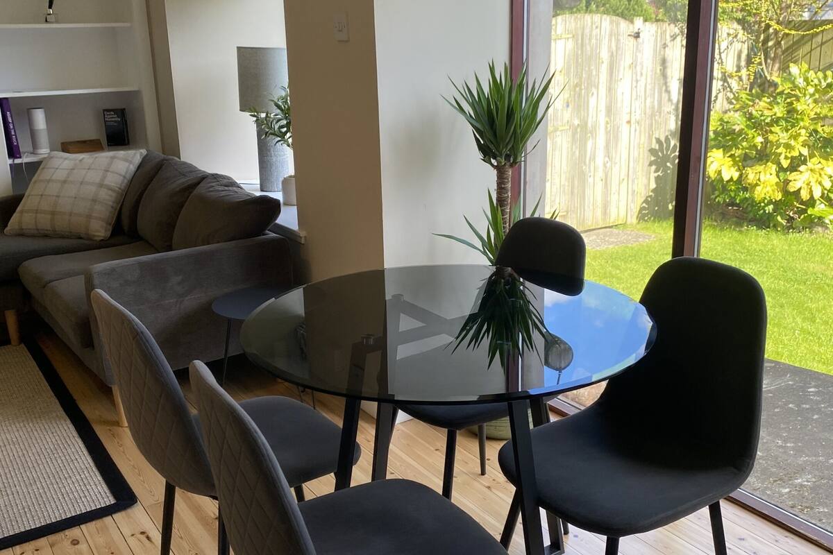 A modern living room with a glass table, four chairs, a sofa, and a large window offering a view of a garden.