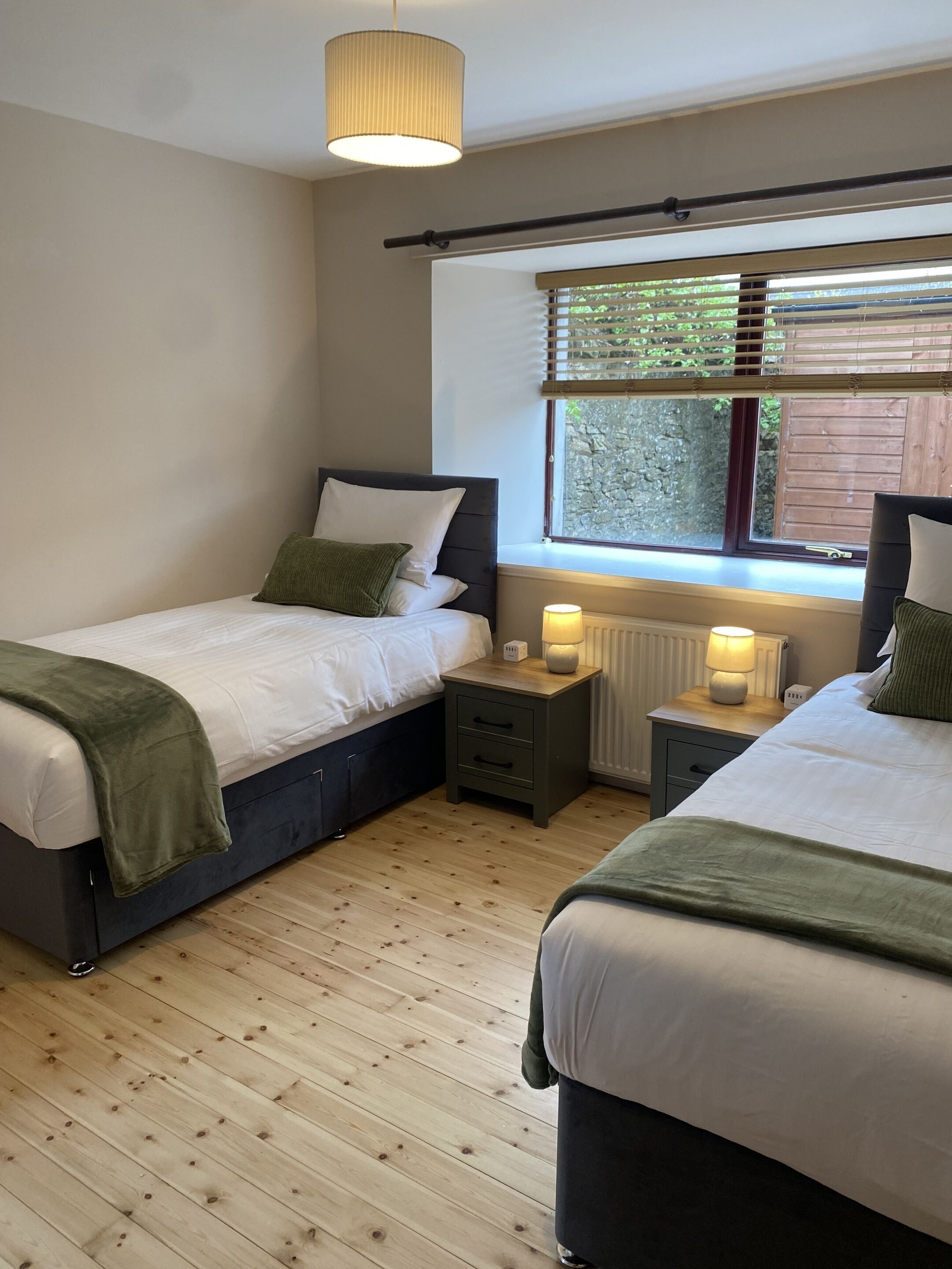 A bedroom with two beds, a window with blinds, a radiator, and a hanging light fixture.