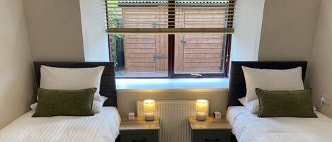 A twin room with two beds, each with a nightstand, a lamp, and a window with blinds.