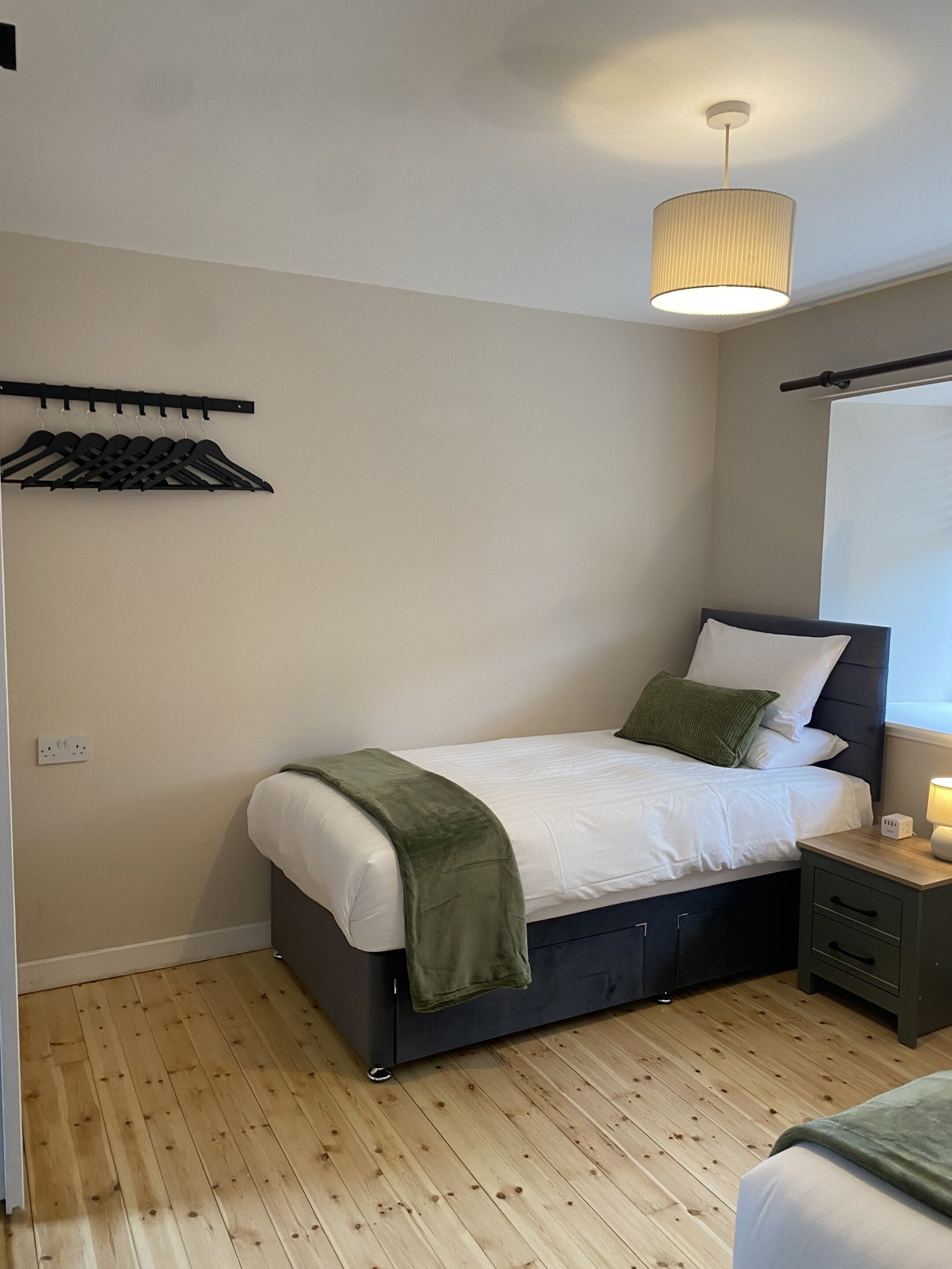 A bedroom with a bed, bedside table, lamp, and a hanging clothes rack.