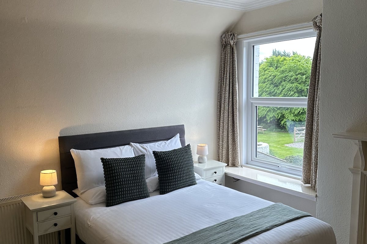 A neatly arranged bedroom with a large bed, bedside lamps, a window with curtains, and a ceiling light fixture.