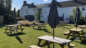A grassy outdoor area with wooden picnic tables and umbrellas, a white building in the background, and a clear blue sky. - Smiddy Haugh (Auchterarder)