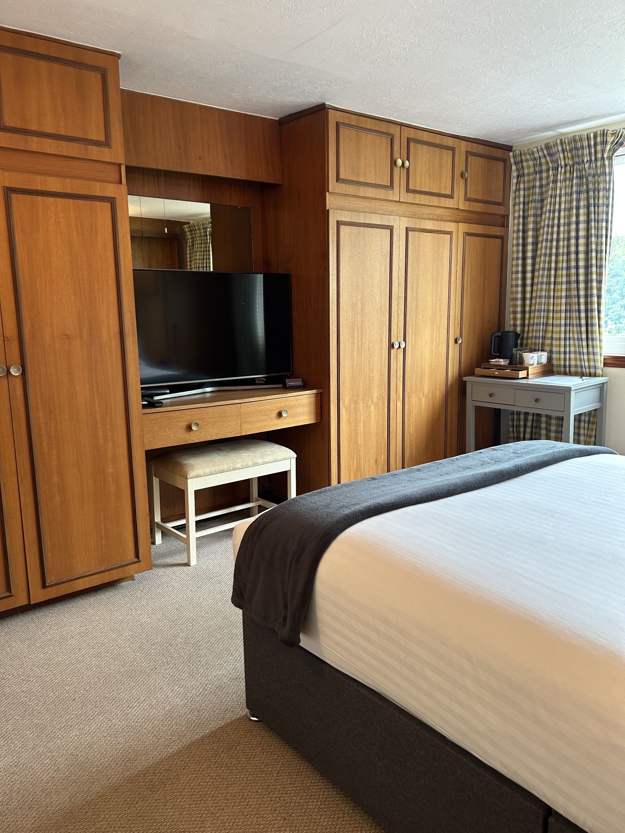 Habitación de hotel con una cama grande, armario de madera, televisor sobre un mueble bajo, una mesita con tetera y una ventana con cortinas a cuadros.