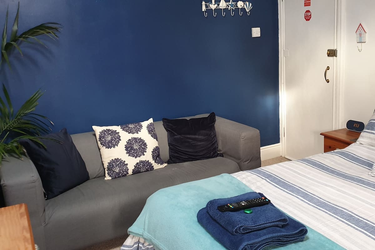A room with a grey sofa, a bed with striped bedding, a blue blanket, and a remote control on the bed.
