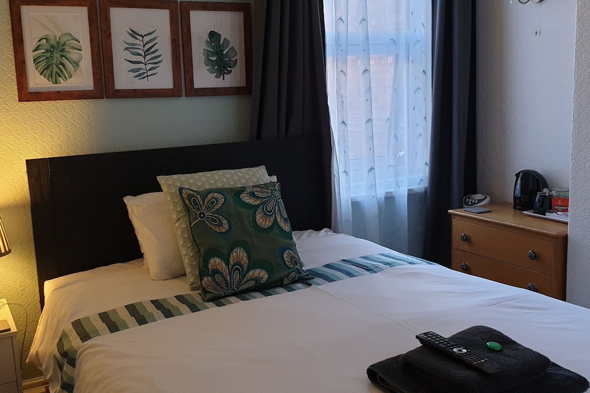 A neatly made bed with a striped pillow and a decorative pillow, a bedside table with a lamp, a nightstand, and a drawer with a clock.