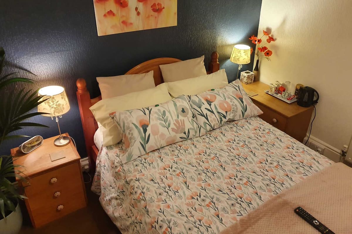 A neatly made bed with floral bedding, a bedside lamp, and a nightstand with a phone and a plant.