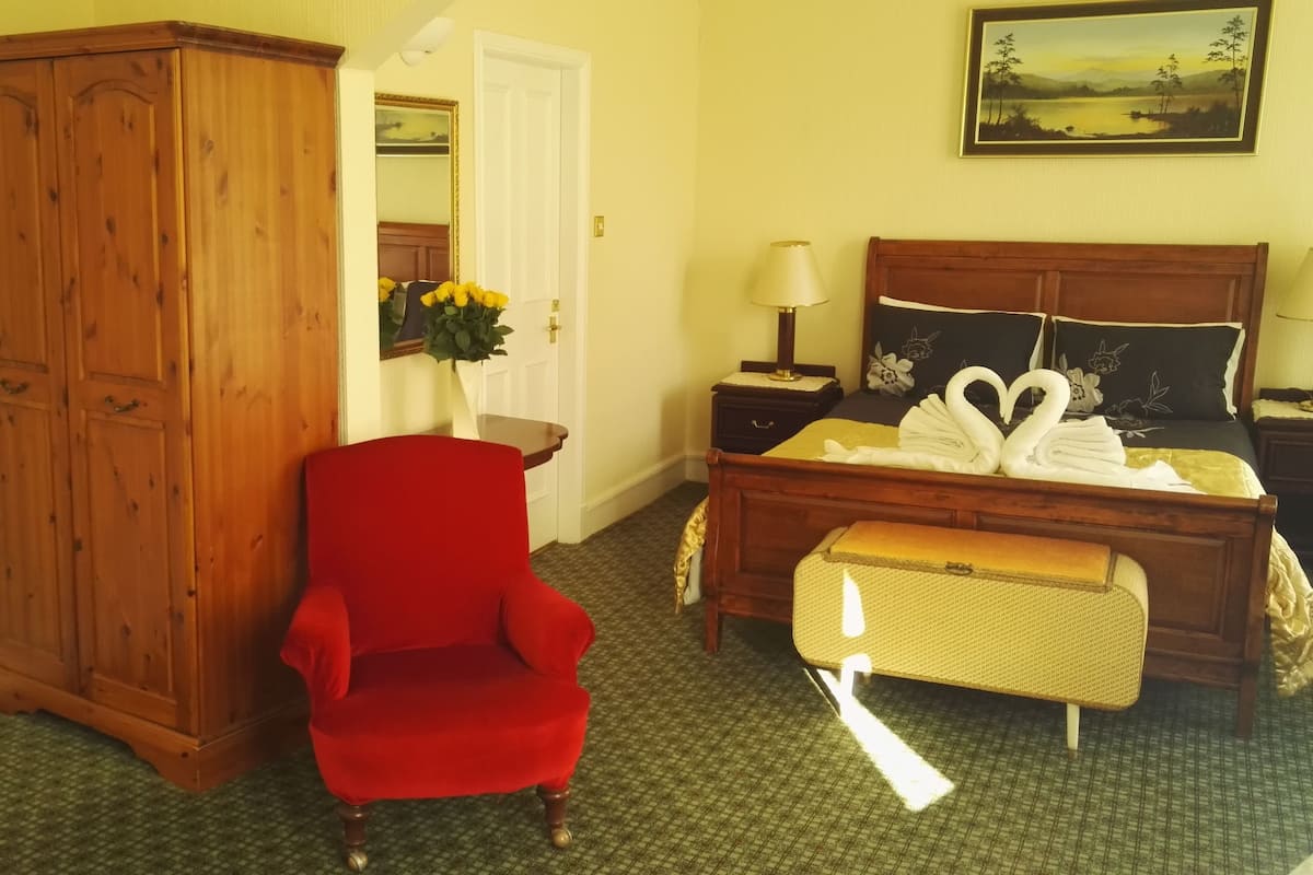 A bedroom with a wooden wardrobe, a red armchair, a bed with a swan-shaped towel, and a painting on the wall.