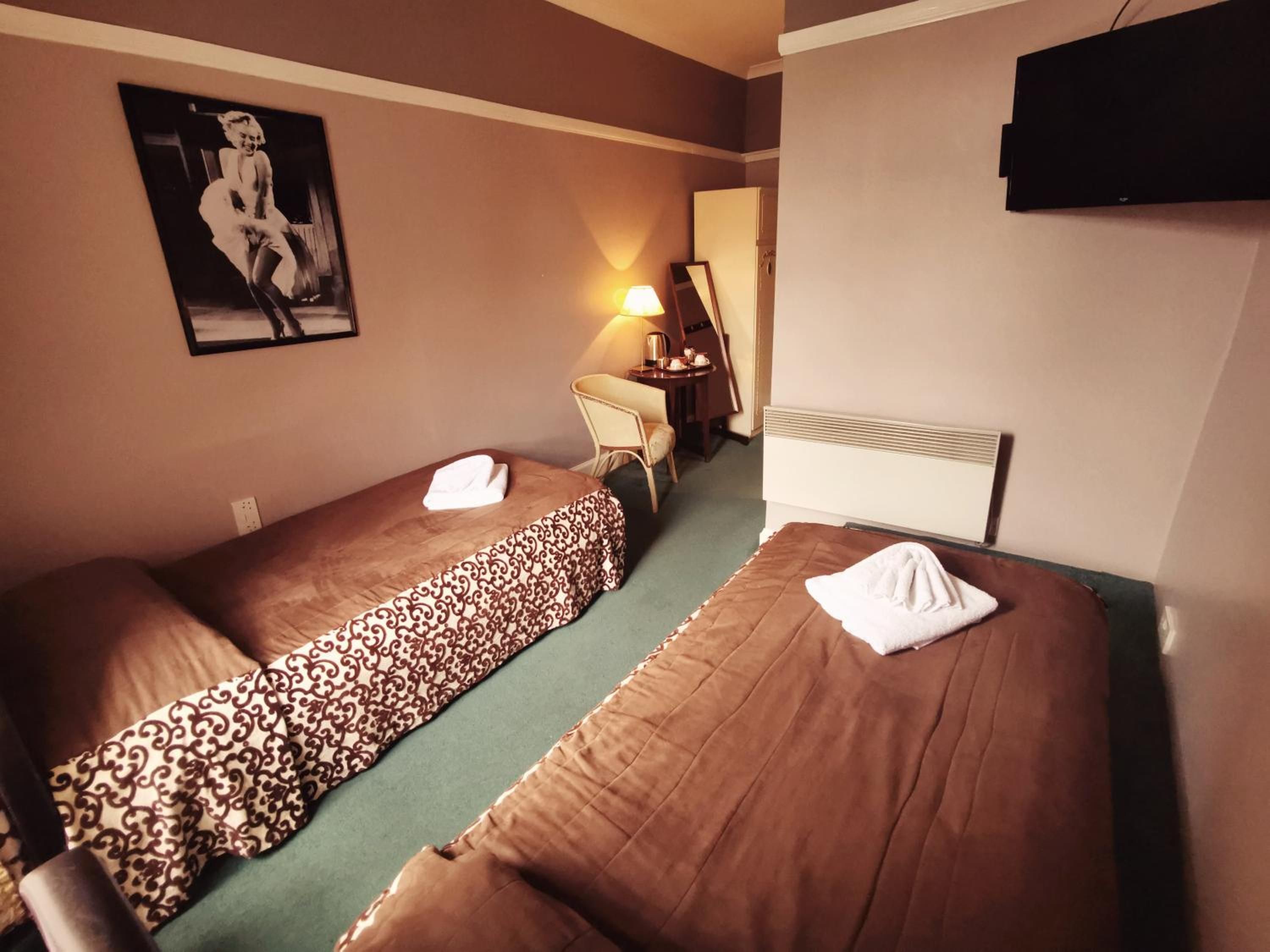 Ein Hotelzimmer mit zwei Einzelbetten, einem Schreibtisch mit Stuhl, einer Lampe, einem Fernseher und einem gerahmten Bild an der Wand.