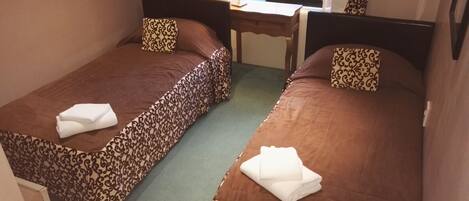 Una habitación pequeña con dos camas, cada una con una colcha estampada y almohadas a juego. Hay un escritorio con una lámpara y una ventana con cortinas.