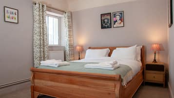A neatly made bed with white linens and a green throw, two bedside lamps, a window with curtains, and two framed pictures on the wall.
