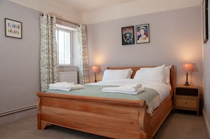 Una cama bien hecha con sábanas blancas y un plaid verde, dos lámparas de noche, una ventana con cortinas y dos cuadros en la pared.