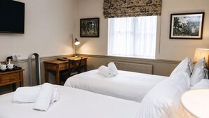 A hotel room with two beds, a desk, a chair, a lamp, and a window with curtains. - George Inn, Mere (Warminster)