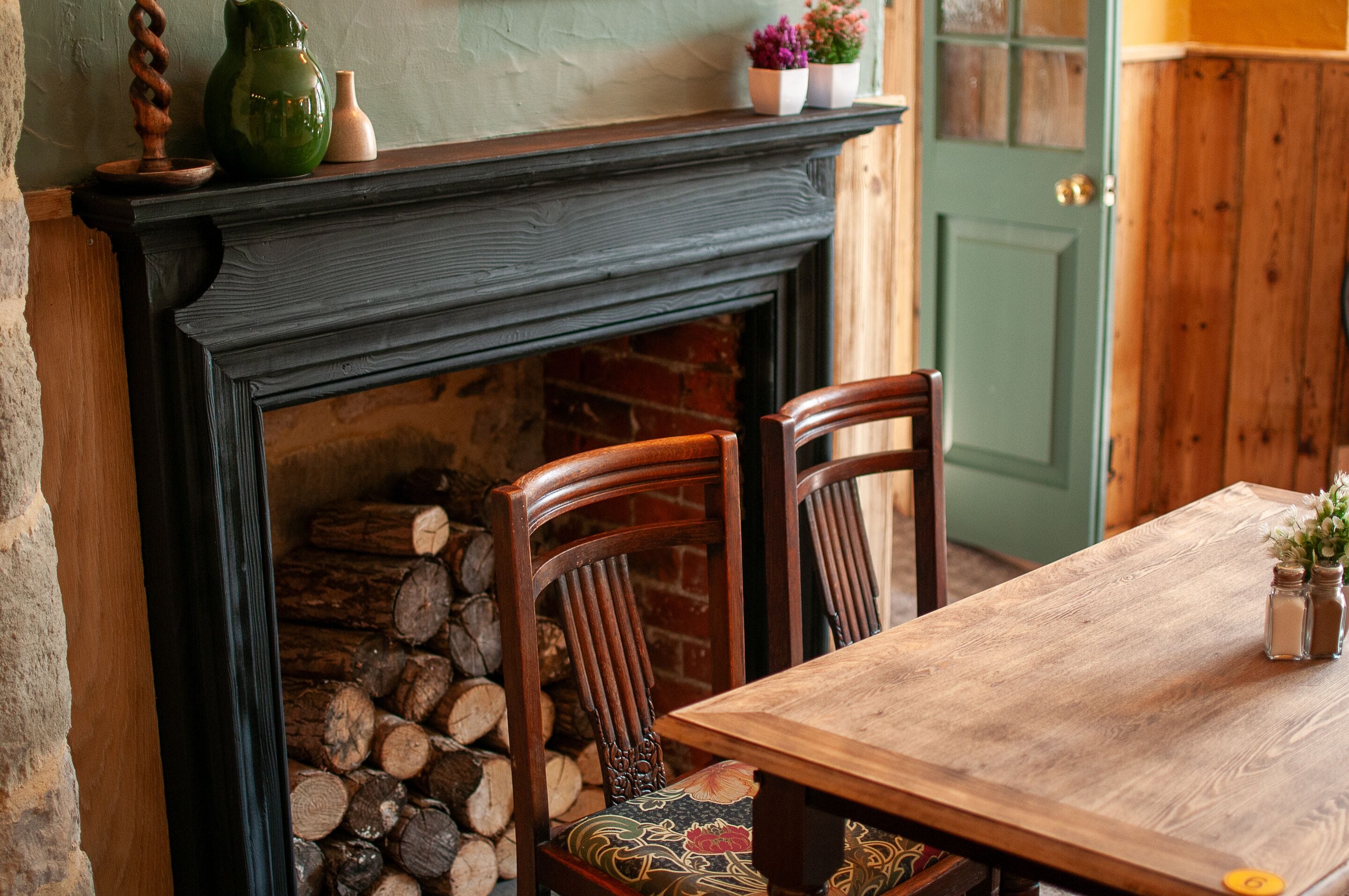 Una chimenea con repisa, una mesa, sillas y una puerta al fondo.