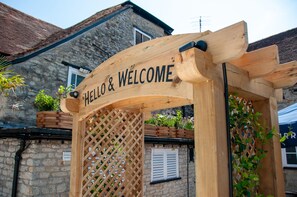A wooden archway with 'HELLO & WELCOME' written on it, leading to a building with stone walls and a tiled roof.