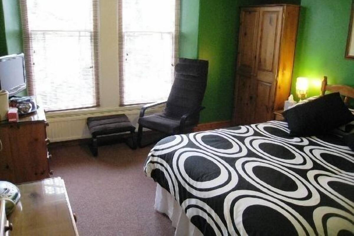 A room with a bed, a chair, a wooden cabinet, and a window with blinds.