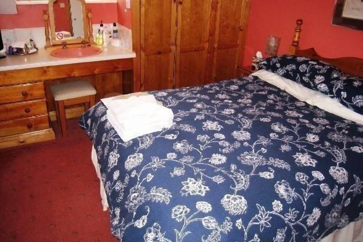 A bedroom with a wooden wardrobe, a bedside table, a mirror, and a bed with a floral bedspread.