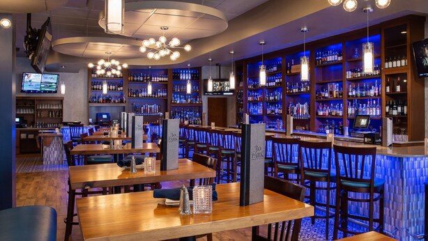 A bar area with wooden tables and chairs, a bar counter with a digital screen, and shelves stocked with bottles. - Hilton Garden Inn Clifton Park (Clifton Park)