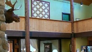A spacious lobby with a high ceiling, wooden walls, and a patterned wall decoration. There are several sofas and a coffee table in the center, with a mounted moose head on the left. - Snowy Owl Inn And Resort (Waterville Valley)