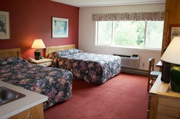 A room with two beds, a small desk, a chair, a window with a view of greenery, and a radiator. - Snowy Owl Inn And Resort (Waterville Valley)