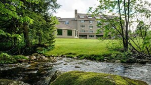 A hotel with a grassy lawn, a stream, and a forested area. - Snowy Owl Inn And Resort (Waterville Valley)