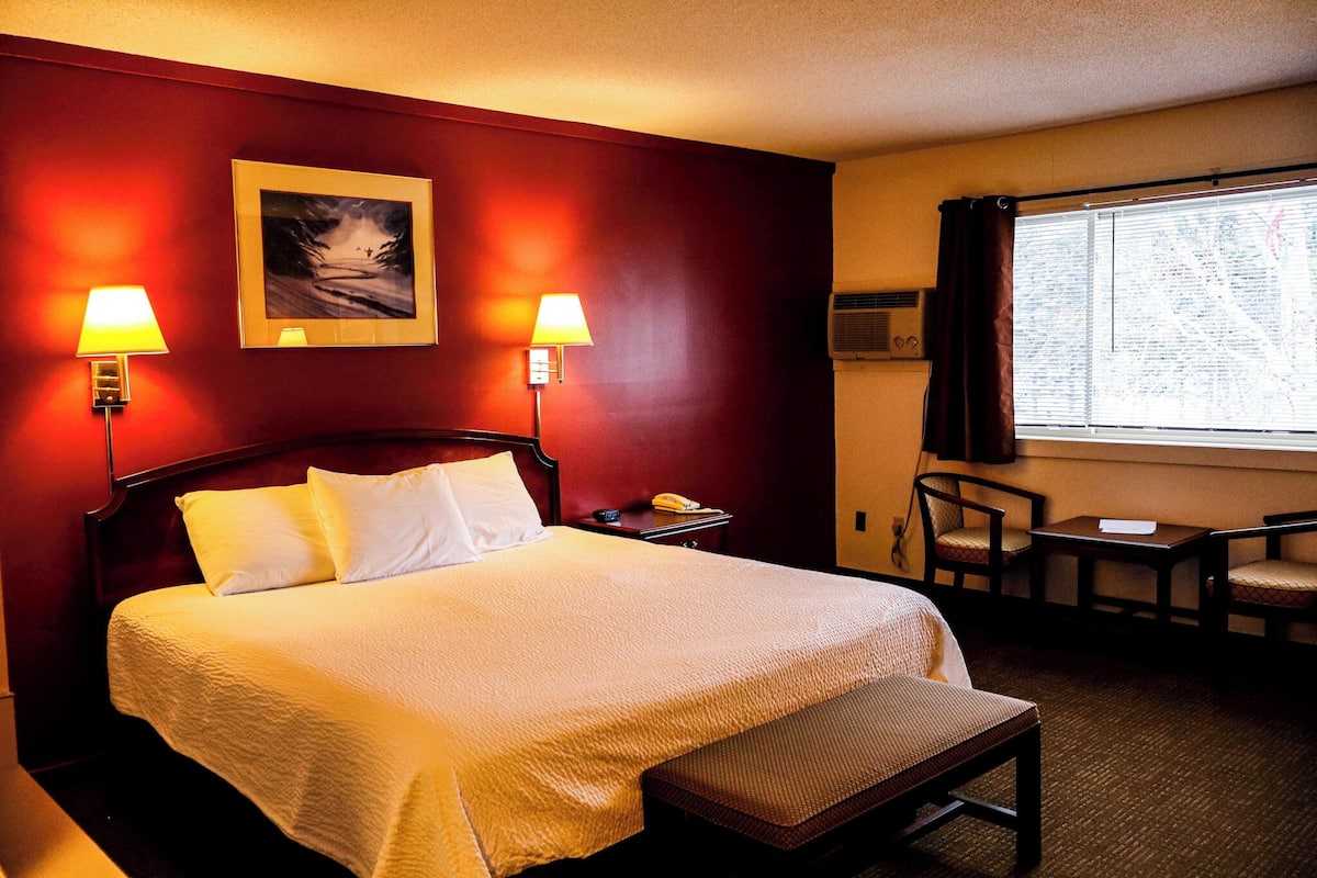 A hotel room with a large bed, two bedside lamps, a framed picture on the wall, a small table with a chair, and a window with curtains.