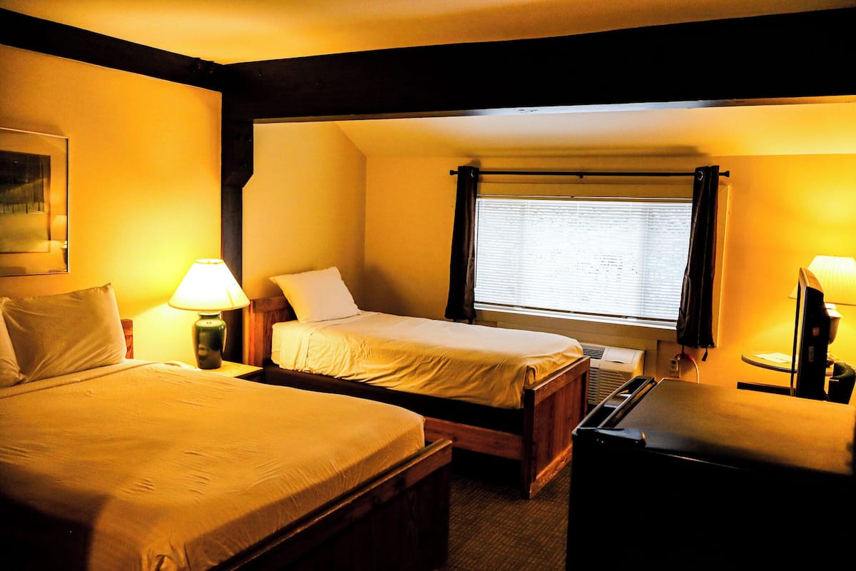 A hotel room with two beds, a desk, a lamp, and a window with blinds.