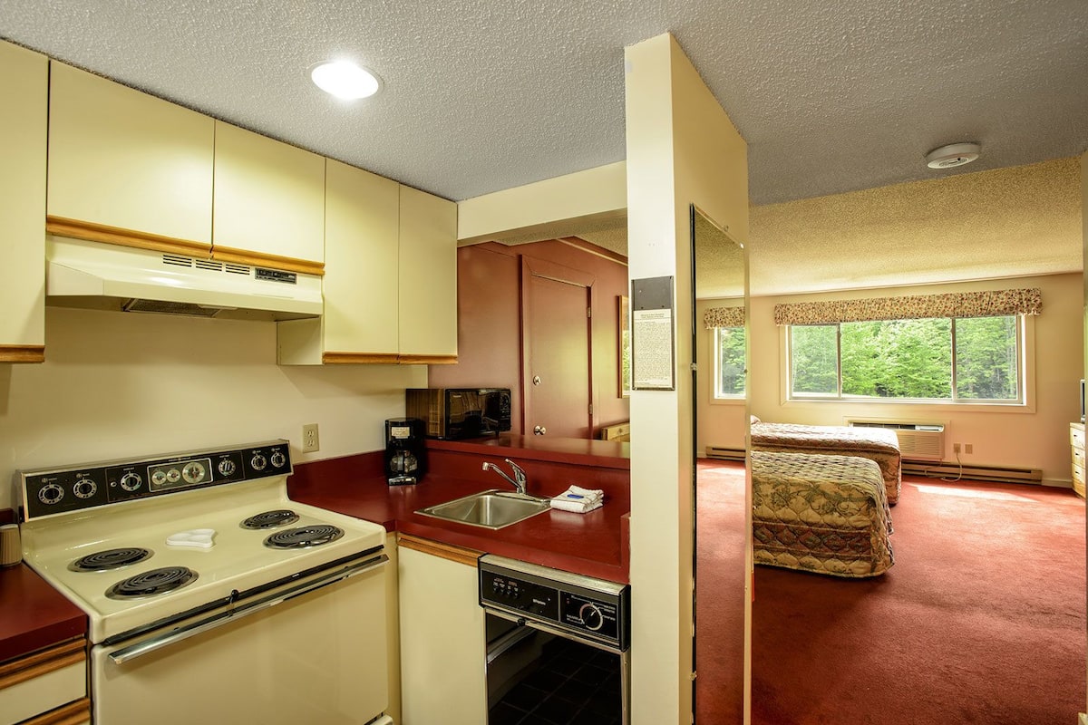 A compact kitchen with a stove, sink, and microwave. There is a small dining area with a table and chairs. The room has a window with a view of trees.