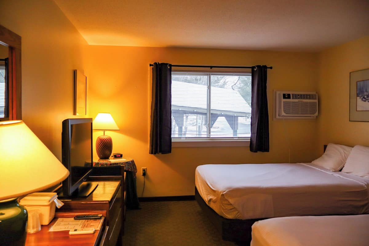 A hotel room with two beds, a desk with a lamp, a window with curtains, an air conditioner, and a framed picture on the wall.