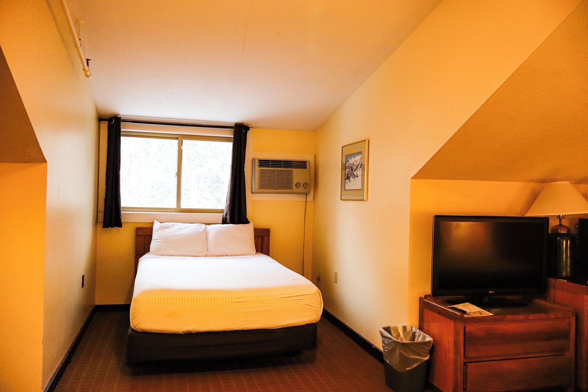 Habitación de hotel con cama, televisor, lámpara, unidad de aire acondicionado y un cuadro en la pared.