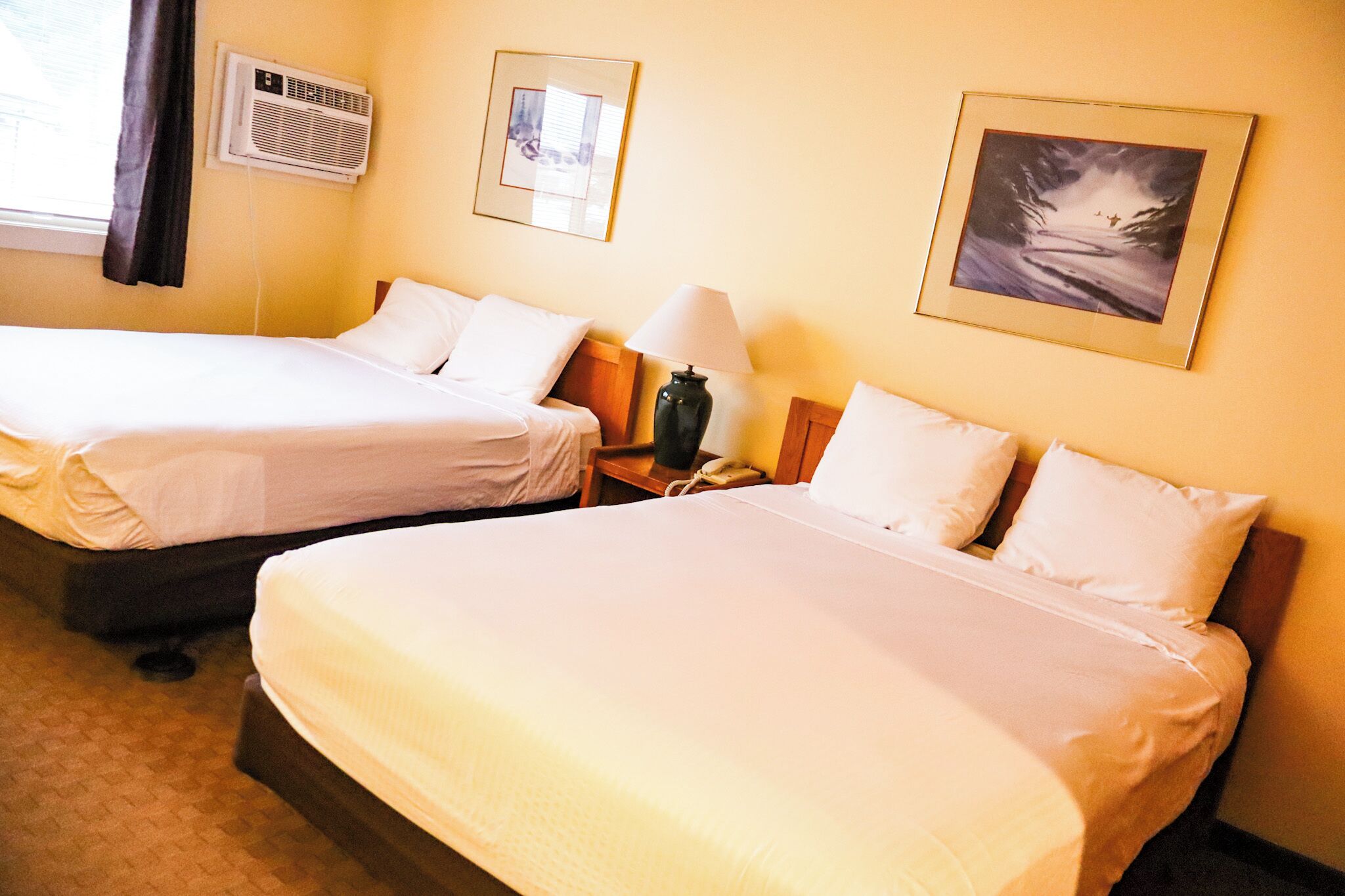 Habitación de hotel con dos camas individuales, una mesita de noche con lámpara, unidad de aire acondicionado y un cuadro enmarcado en la pared.