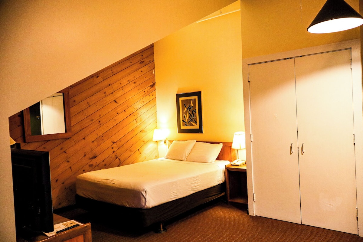 A bedroom with a wooden sloped ceiling, a bed with white linens, two bedside lamps, a framed picture on the wall, and a wardrobe.