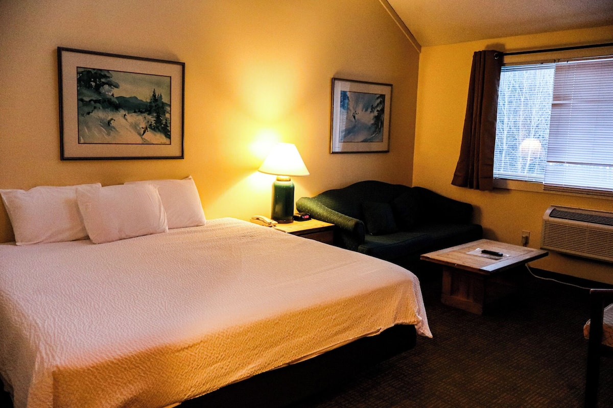A hotel room with a bed, a desk, a chair, a lamp, and two framed pictures on the wall.