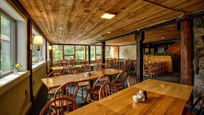 A spacious dining area with wooden tables and chairs, large windows, and a stone wall. - Snowy Owl Inn And Resort (Waterville Valley)