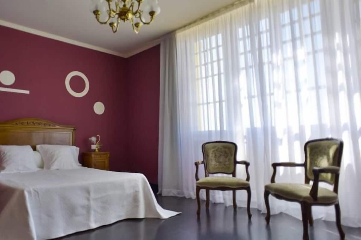 A bedroom with a large bed, two nightstands, a chandelier, and two upholstered chairs.