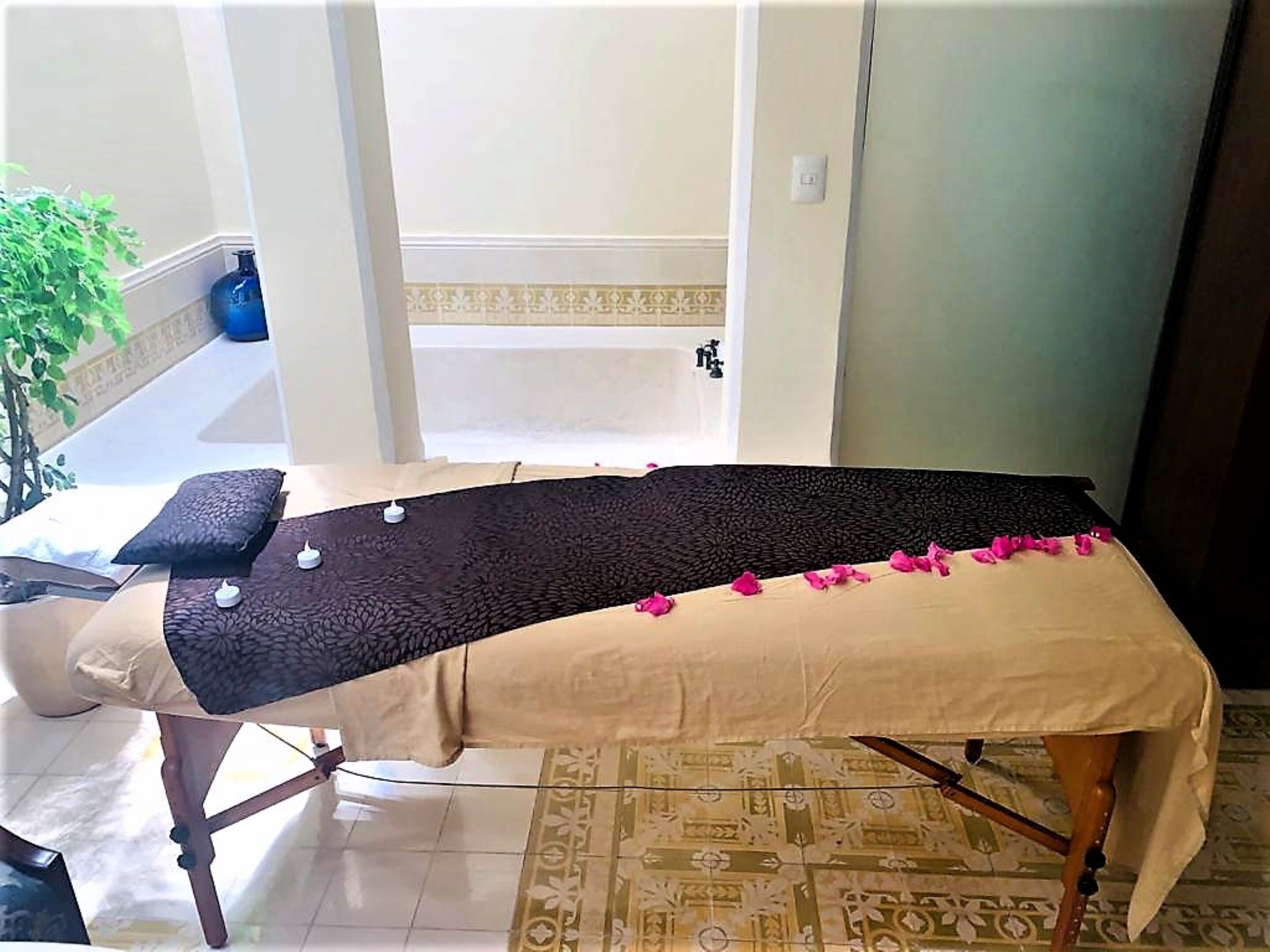 A massage table with a patterned black mat and beige cover, surrounded by a patterned floor and a small plant.