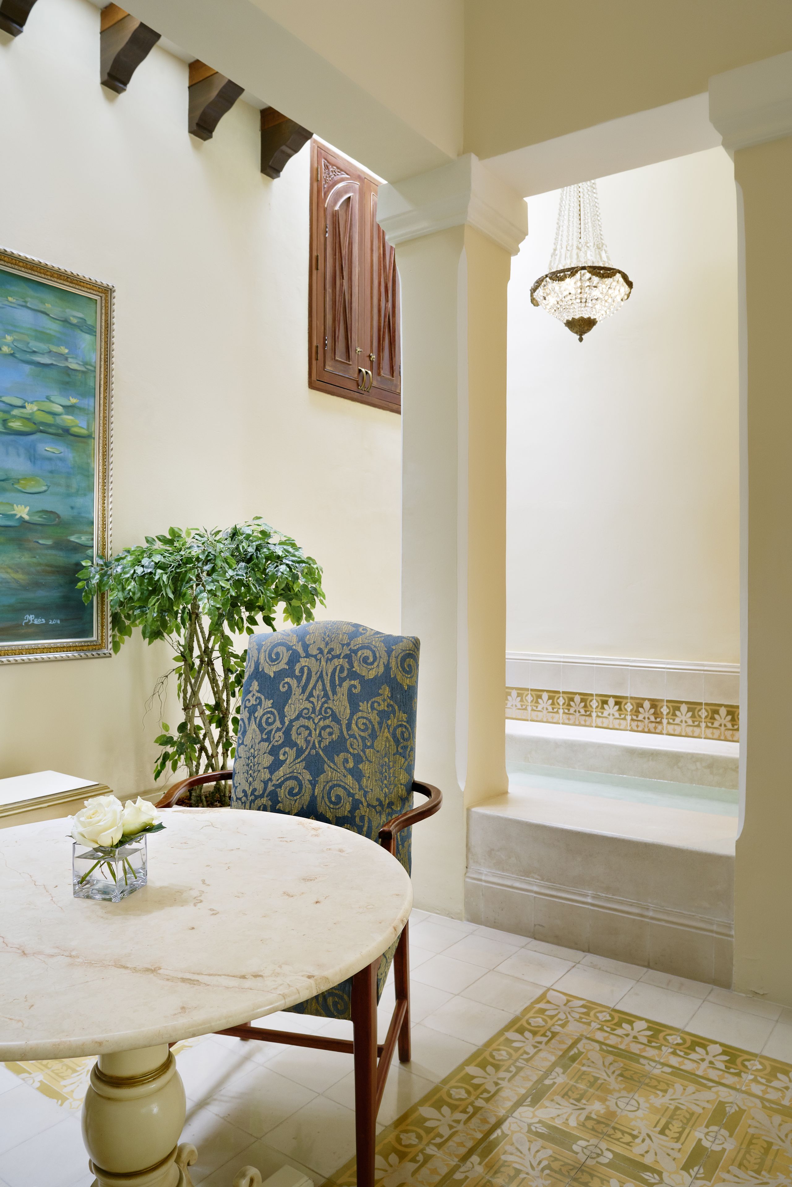 A room with a round table, a chair, a potted plant, a painting, and a view of a pool.