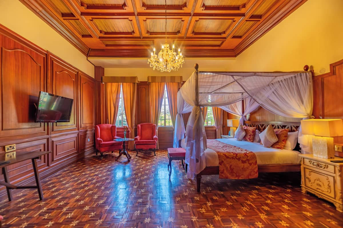 Quarto com piso de madeira, cama com dossel, televisão, poltrona vermelha e lustre.