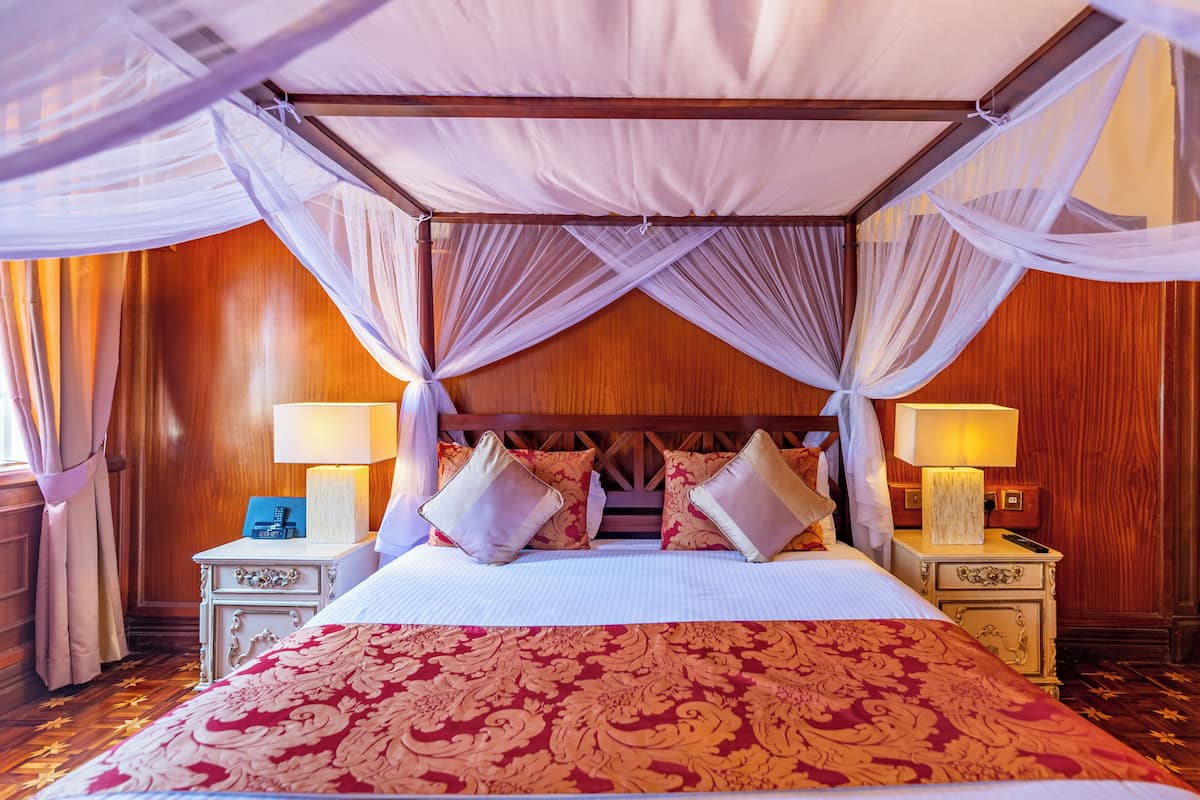 Uma cama de dossel com estrutura de madeira, duas mesas de cabeceira com abajures e um cobreleito vermelho estampado.