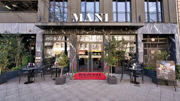 Un edificio con el letrero 'MANI Restaurant', terraza con mesas y sillas, y entrada con alfombra roja.