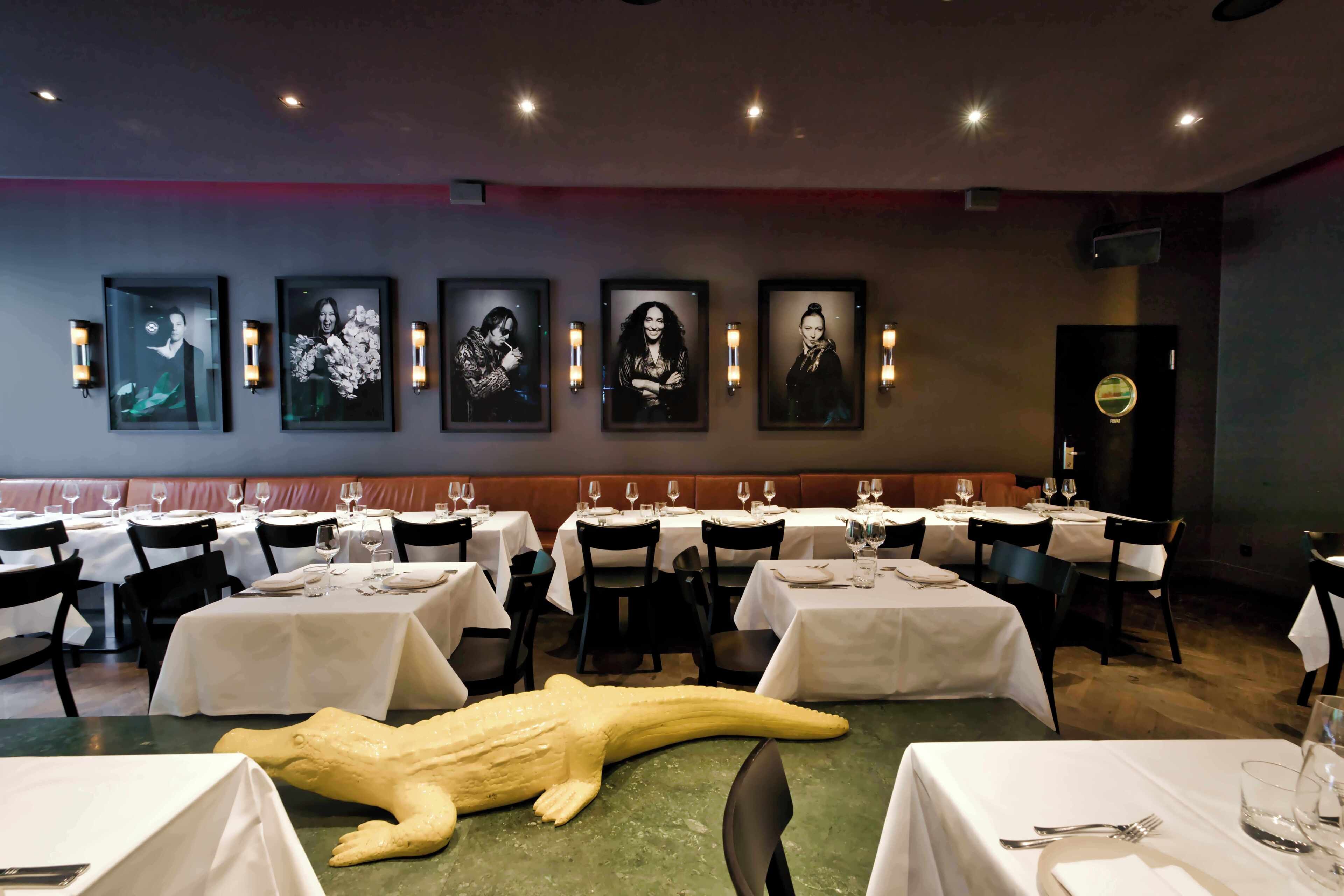 A restaurant with tables set for dining, framed portraits on the wall, and a large sculpture on the floor.